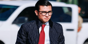 Man Wearing Glasses, White Shirt, Red Tie and Quilted Jacket Walking Outdoors with a White Suv Behind Him. - Salud Regia