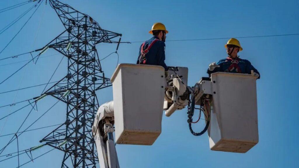 Two Utility Workers in Yellow Hard Hats Stand in Elevated Bucket Baskets, Inspecting Power Lines Against a Clear Blue Sky, Connected by Safety Harnesses. - Salud Regia