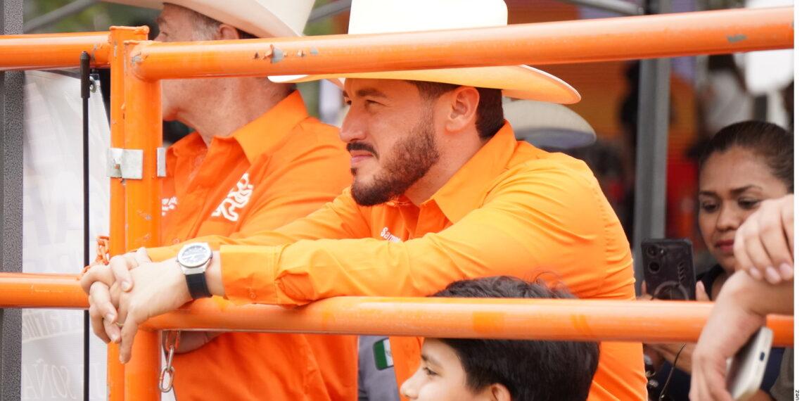 Man in an Orange Shirt and White Cowboy Hat Leans on an Orange Barricade at a Crowded Outdoor Event. - Salud Regia