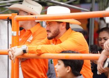 Man in an Orange Shirt and White Cowboy Hat Leans on an Orange Barricade at a Crowded Outdoor Event. - Salud Regia