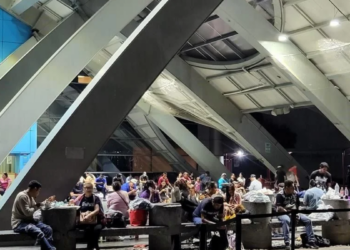 Crowded Indoor Waiting Area with People Sitting at Tables and Benches Beneath Large Diagonal Steel Beams. - Salud Regia