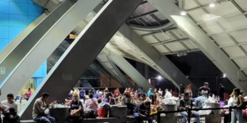 Crowded Indoor Waiting Area with People Sitting at Tables and Benches Beneath Large Diagonal Steel Beams. - Salud Regia