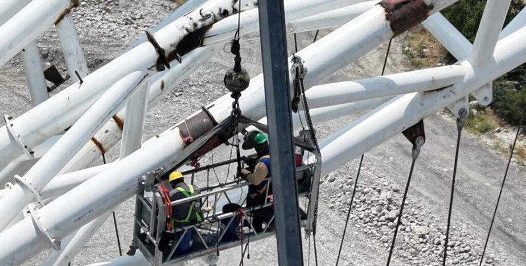 Maintenance Crew in a Suspended Basket Working on a Tall White Steel Tower Frame, Wearing Hard Hats and Safety Gear. - Salud Regia